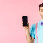 E-commerce and shopping concept. Funny guy look curious at empty smartphone screen, showing money, buying online, standing on pink background