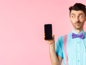 E-commerce and shopping concept. Funny guy look curious at empty smartphone screen, showing money, buying online, standing on pink background